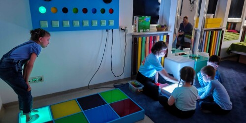 Nauczyciele i dzieci siedzące na dywanie, dziewczynka stojąca na kasetonie dźwiękowym