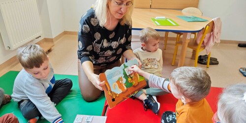 Chłopiec wkleja symbol PCS do książki. Dzieci i nauczycielka siedzą na materacu w kole