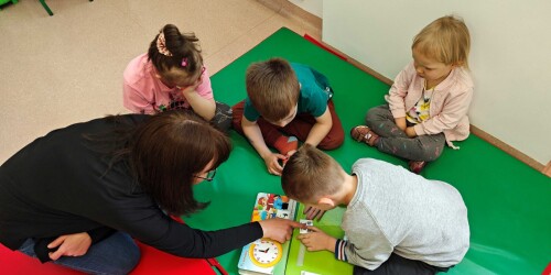 Dzieci wraz ze specjalistą siedzą na dywanie i zaznaczają prawidłowy symbol PCS w książce do czytania uczestniczącego