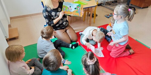 Dzieci siedzą na dywanie i słuchają czytanej książki przez nauczycielkę